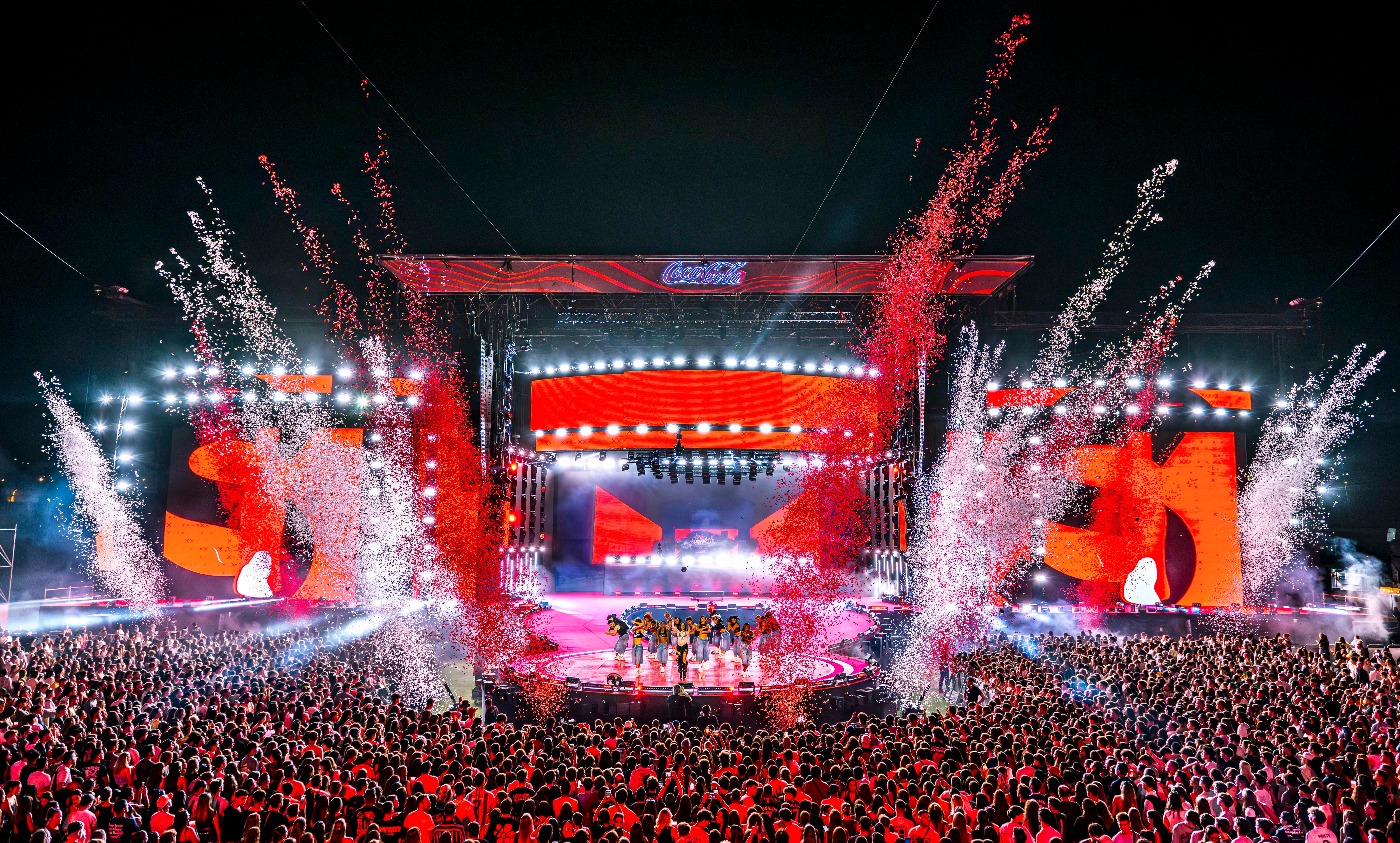 Spektakuläres Musikfestival bei Nacht mit einer großen rot beleuchteten Bühne, einer riesigen Menschenmenge und einer Explosion aus rotem und weißem Konfetti.
